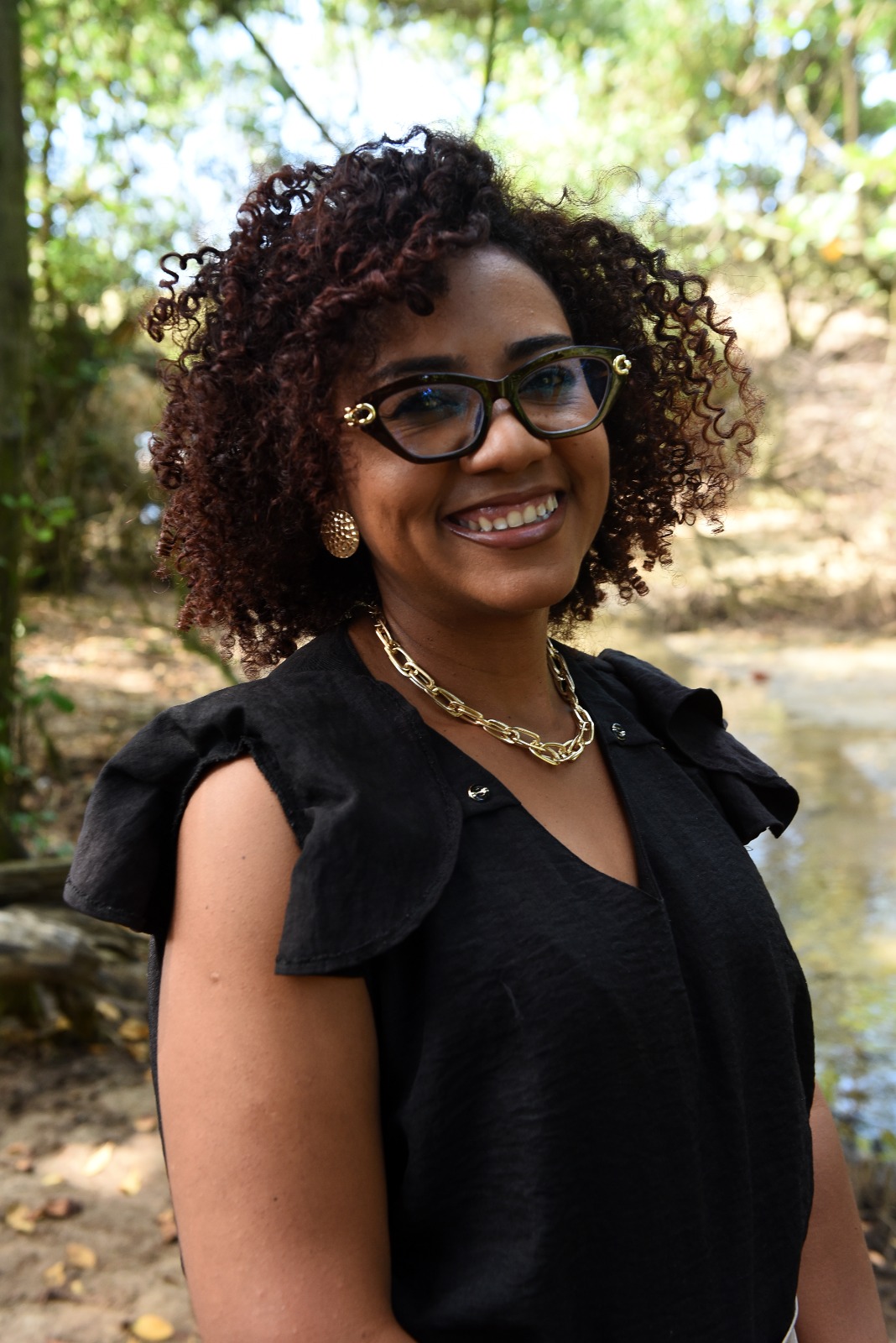 Retrato de Priscila Santos sorrindo, de óculos e blusa preta, ao ar livre