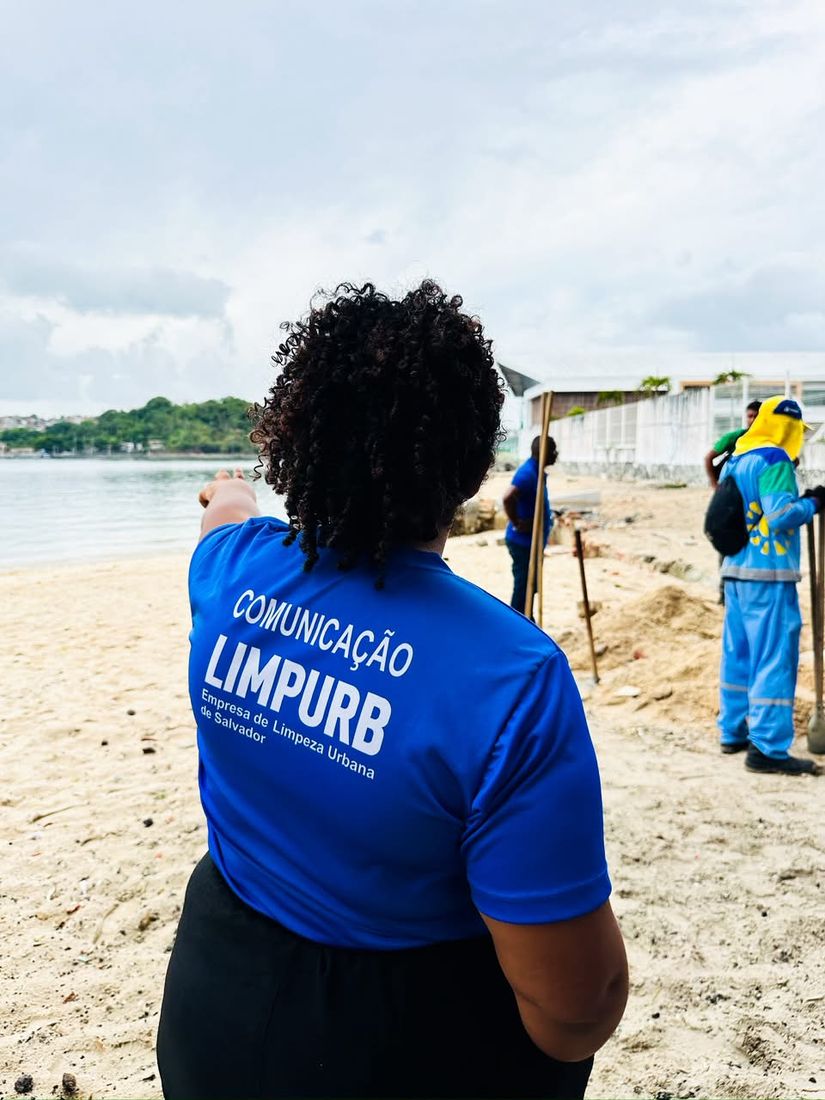 Priscila de costas, com a camisa Comunicação Limpurb, orientando uma gravação na praia